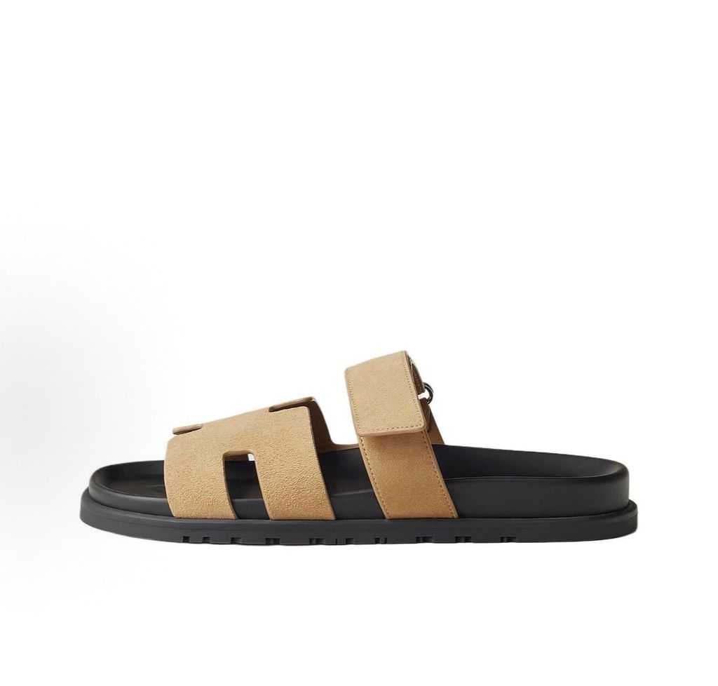 Angled top-down view of the Hermès Genius Sandals in taupe grey suede, showcasing the signature 'H' cutout front strap, the adjustable hook-and-loop instep strap, and the contoured black ergonomic rubber footbed with Hermès branding.