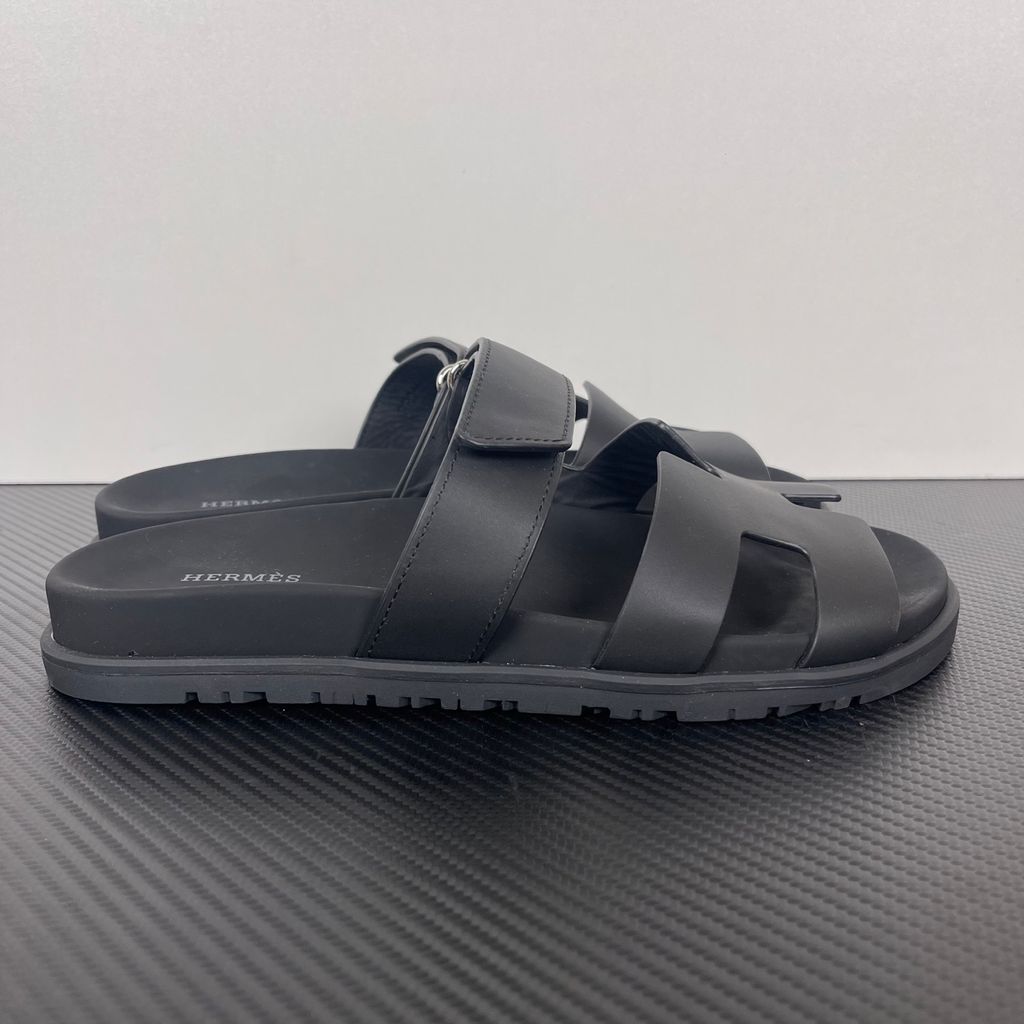 Side profile of the Hermès Genius Sandals in sleek black smooth leather, showcasing the signature 'H' cutout front strap, the adjustable hook-and-loop instep strap, and the contoured black ergonomic rubber footbed with subtle Hermès branding.