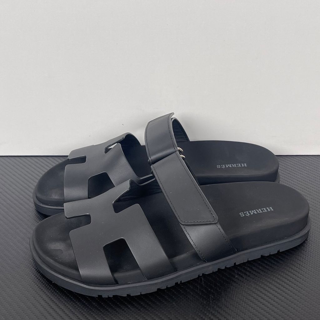 Side profile of the Hermès Genius Sandals in sleek black smooth leather, showcasing the signature 'H' cutout front strap, the adjustable hook-and-loop instep strap, and the contoured black ergonomic rubber footbed with subtle Hermès branding.