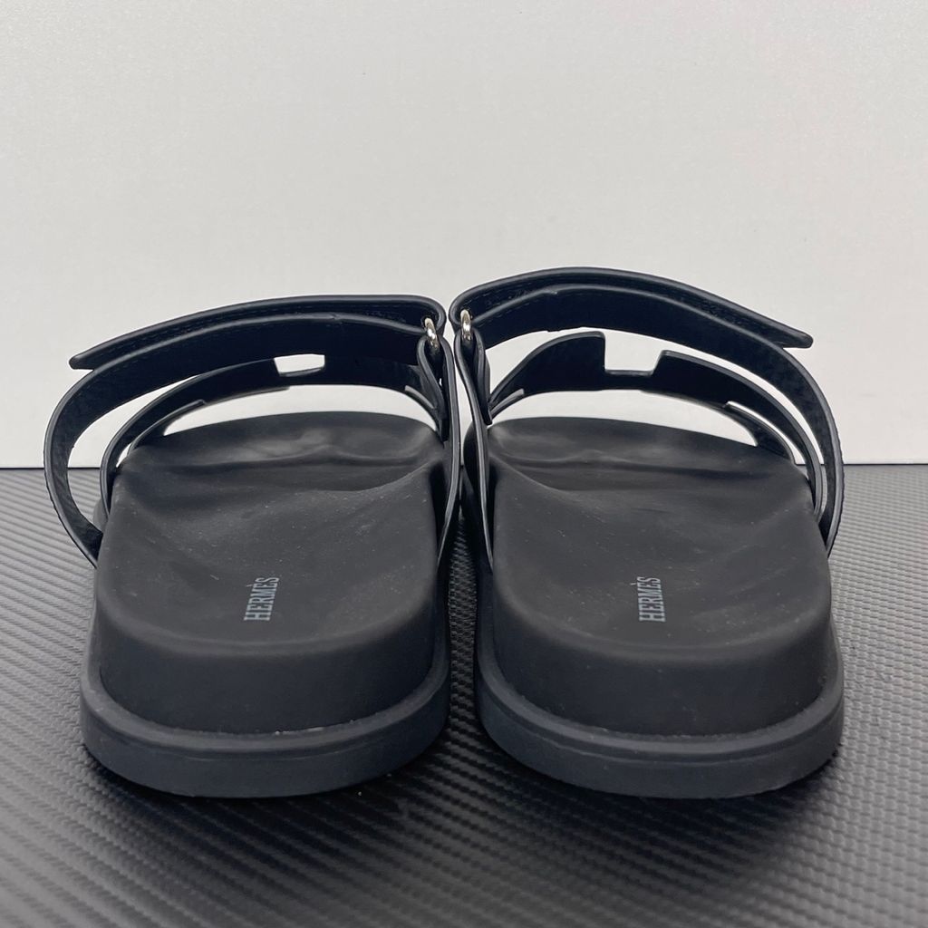 Side profile of the Hermès Genius Sandals in sleek black smooth leather, showcasing the signature 'H' cutout front strap, the adjustable hook-and-loop instep strap, and the contoured black ergonomic rubber footbed with subtle Hermès branding.
