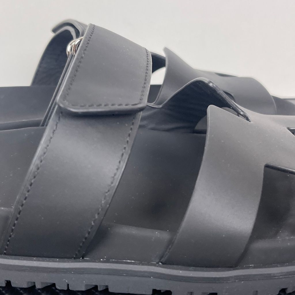 Side profile of the Hermès Genius Sandals in sleek black smooth leather, showcasing the signature 'H' cutout front strap, the adjustable hook-and-loop instep strap, and the contoured black ergonomic rubber footbed with subtle Hermès branding.