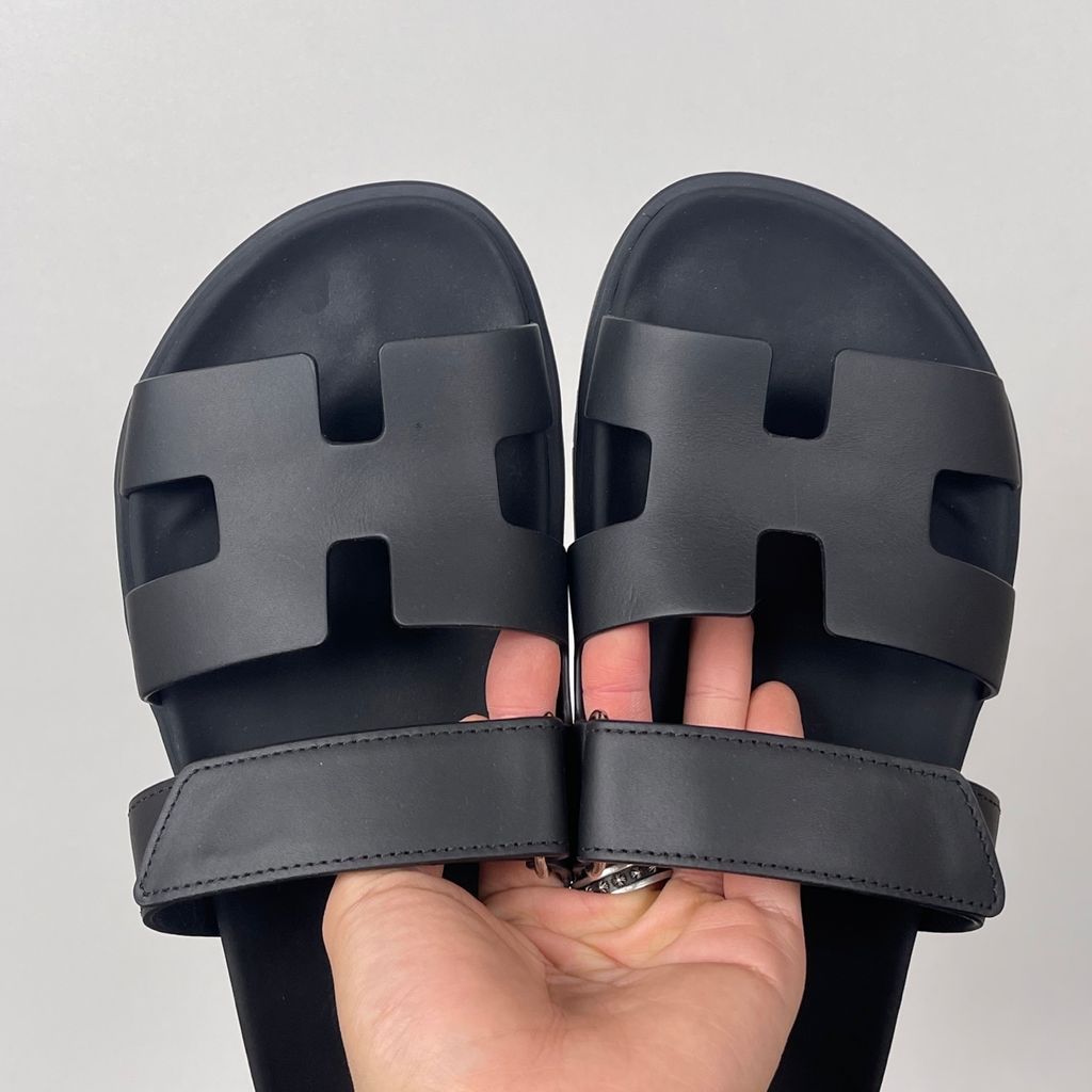 Side profile of the Hermès Genius Sandals in sleek black smooth leather, showcasing the signature 'H' cutout front strap, the adjustable hook-and-loop instep strap, and the contoured black ergonomic rubber footbed with subtle Hermès branding.