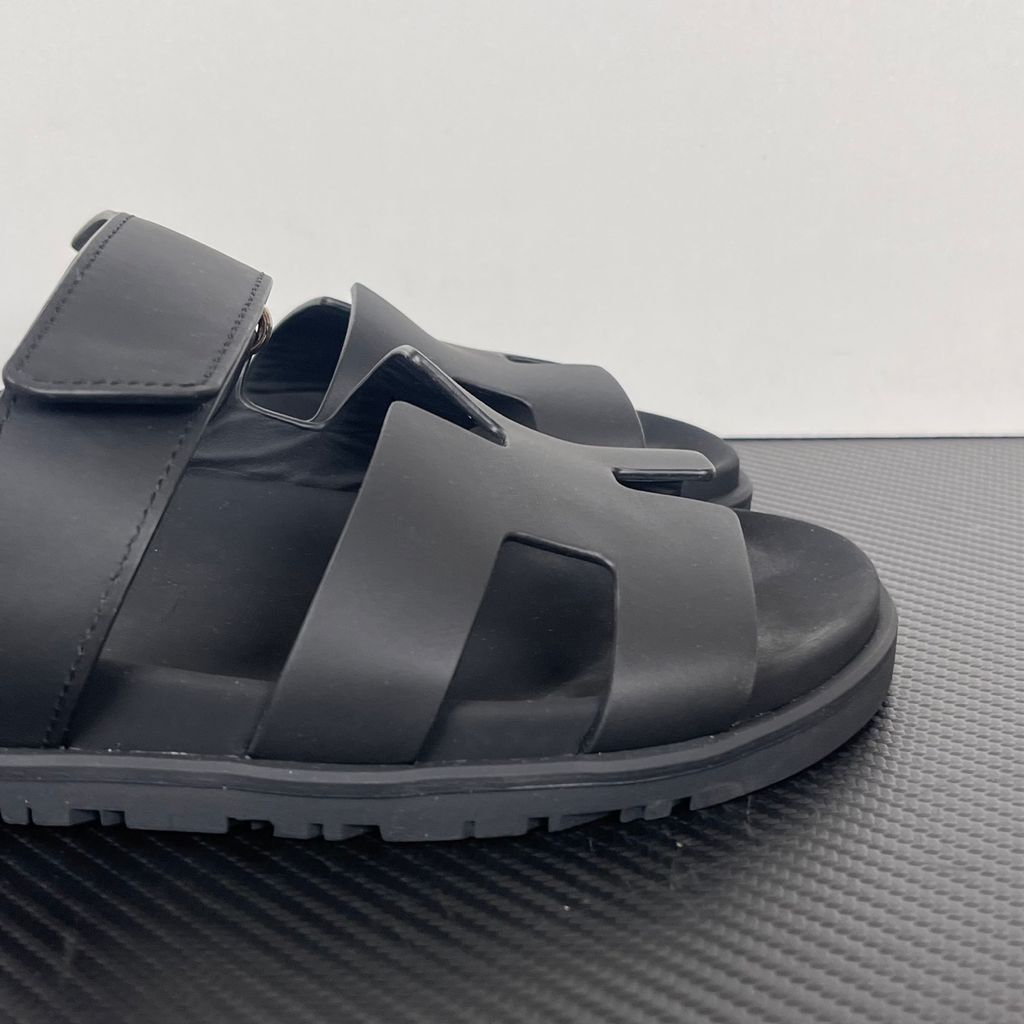 Side profile of the Hermès Genius Sandals in sleek black smooth leather, showcasing the signature 'H' cutout front strap, the adjustable hook-and-loop instep strap, and the contoured black ergonomic rubber footbed with subtle Hermès branding.