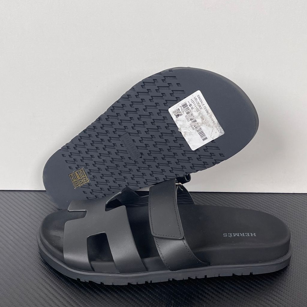 Side profile of the Hermès Genius Sandals in sleek black smooth leather, showcasing the signature 'H' cutout front strap, the adjustable hook-and-loop instep strap, and the contoured black ergonomic rubber footbed with subtle Hermès branding.