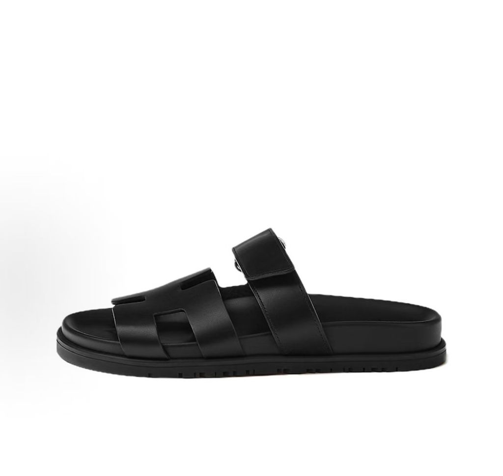 Side profile of the Hermès Genius Sandals in sleek black smooth leather, showcasing the signature 'H' cutout front strap, the adjustable hook-and-loop instep strap, and the contoured black ergonomic rubber footbed with subtle Hermès branding.