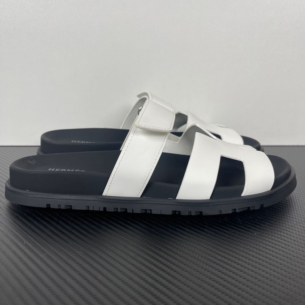 Side profile of the Hermès Genius Sandals in crisp white smooth leather, showcasing the signature 'H' cutout front strap, the adjustable hook-and-loop instep strap, and the contoured black ergonomic rubber footbed with subtle Hermès branding.