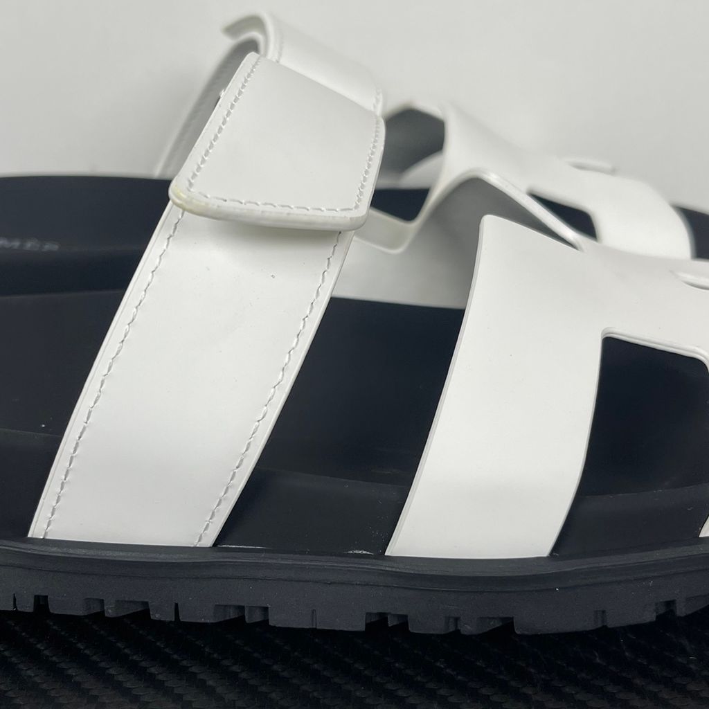 Side profile of the Hermès Genius Sandals in crisp white smooth leather, showcasing the signature 'H' cutout front strap, the adjustable hook-and-loop instep strap, and the contoured black ergonomic rubber footbed with subtle Hermès branding.