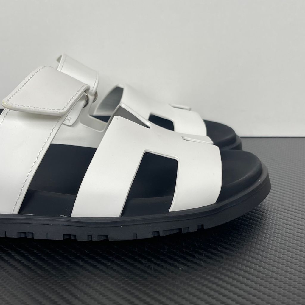 Side profile of the Hermès Genius Sandals in crisp white smooth leather, showcasing the signature 'H' cutout front strap, the adjustable hook-and-loop instep strap, and the contoured black ergonomic rubber footbed with subtle Hermès branding.