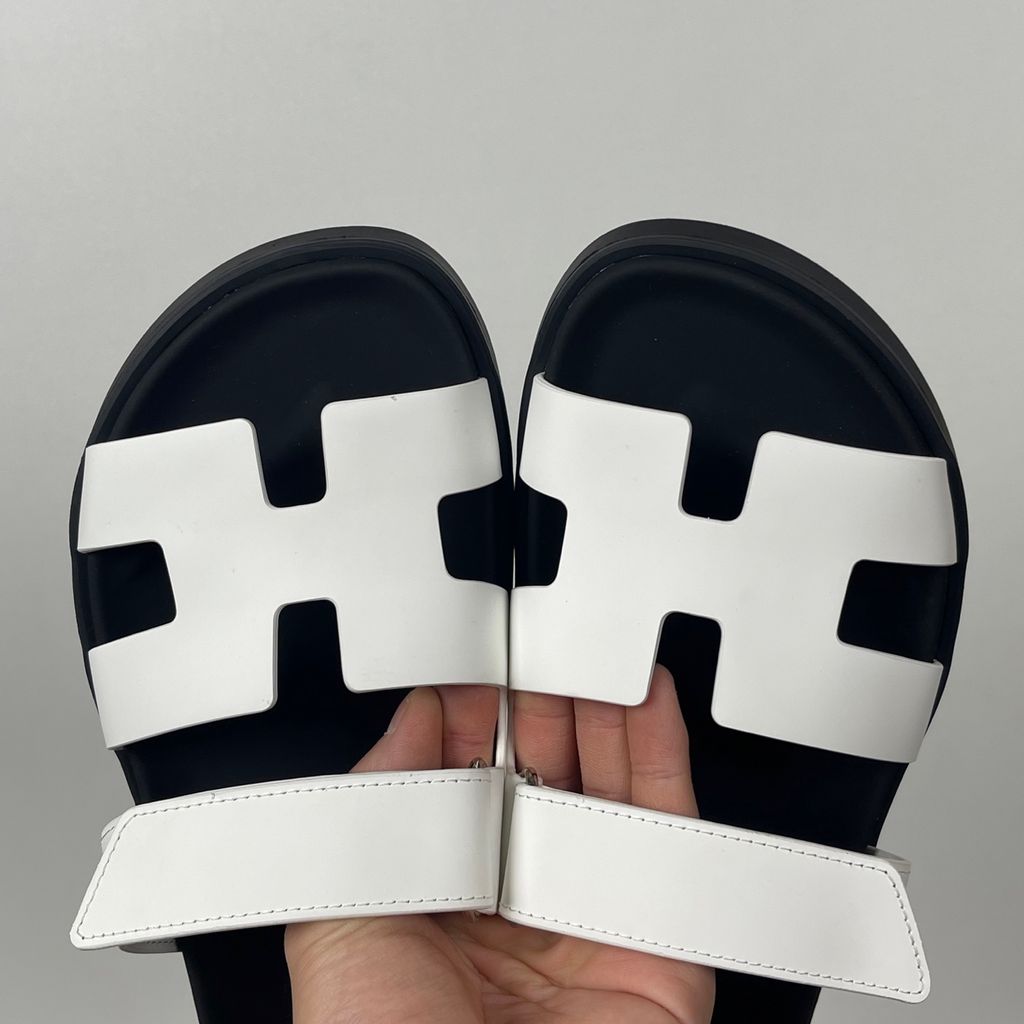 Side profile of the Hermès Genius Sandals in crisp white smooth leather, showcasing the signature 'H' cutout front strap, the adjustable hook-and-loop instep strap, and the contoured black ergonomic rubber footbed with subtle Hermès branding.
