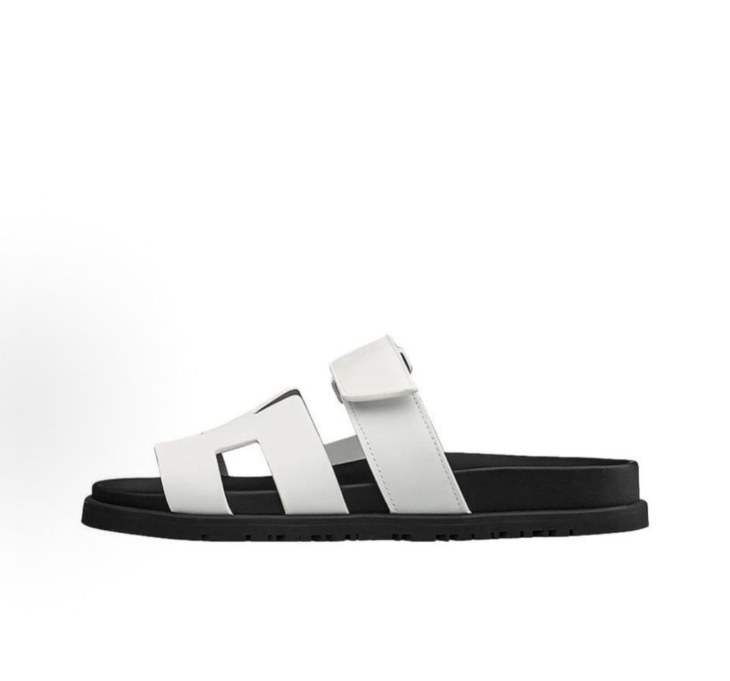 Side profile of the Hermès Genius Sandals in crisp white smooth leather, showcasing the signature 'H' cutout front strap, the adjustable hook-and-loop instep strap, and the contoured black ergonomic rubber footbed with subtle Hermès branding.