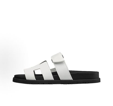 Side profile of the Hermès Genius Sandals in crisp white smooth leather, showcasing the signature 'H' cutout front strap, the adjustable hook-and-loop instep strap, and the contoured black ergonomic rubber footbed with subtle Hermès branding.