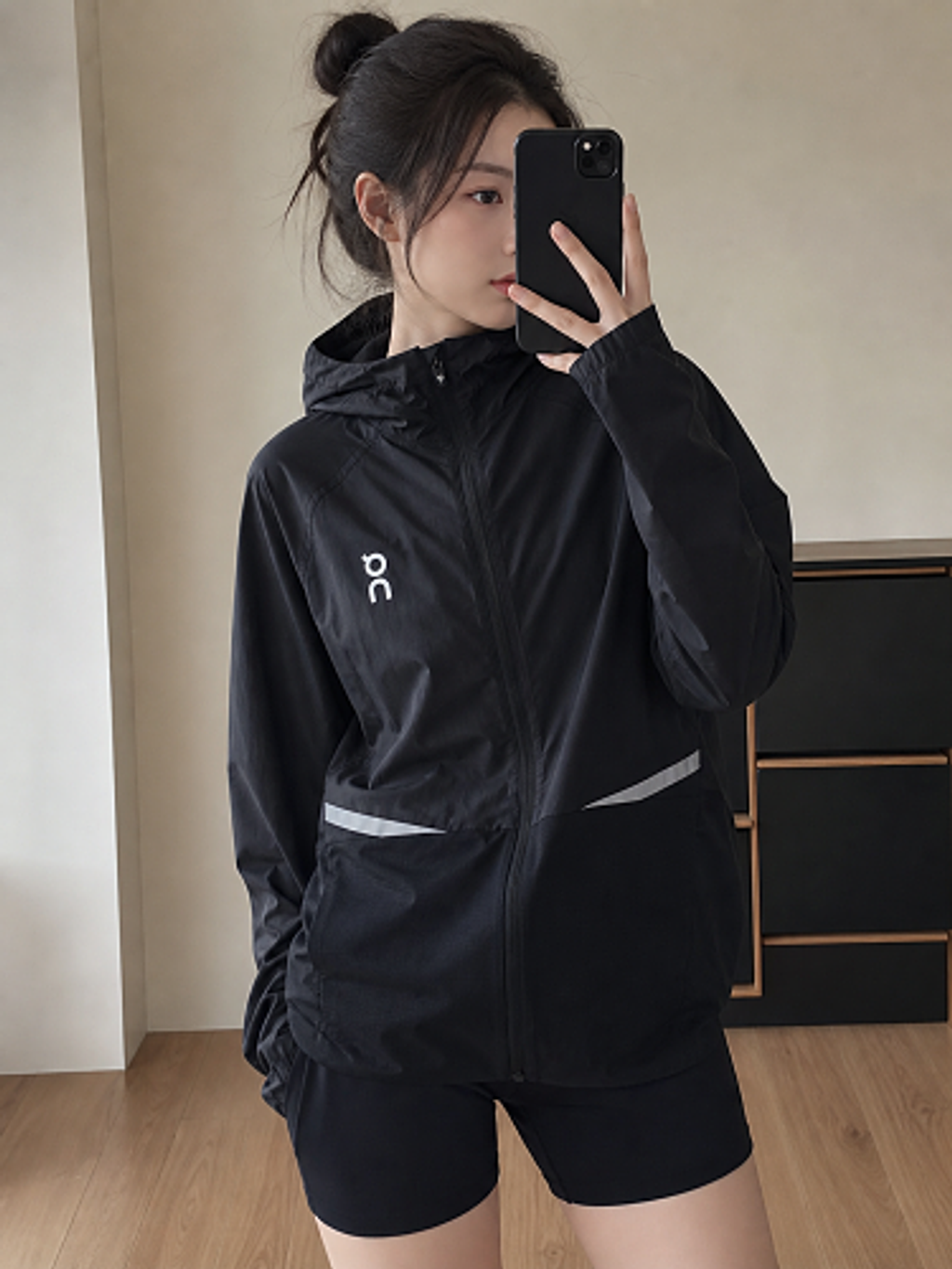 Front profile of the men's On Running Core Jacket in black, showcasing the lightweight windproof fabric, full-length front zipper, protective hood, white 'On' logo, and sleek reflective strips across the midsection for night safety.