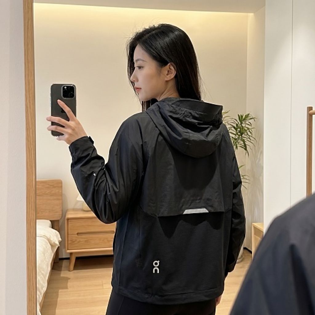 Front profile of the men's On Running Core Jacket in black, showcasing the lightweight windproof fabric, full-length front zipper, protective hood, white 'On' logo, and sleek reflective strips across the midsection for night safety.