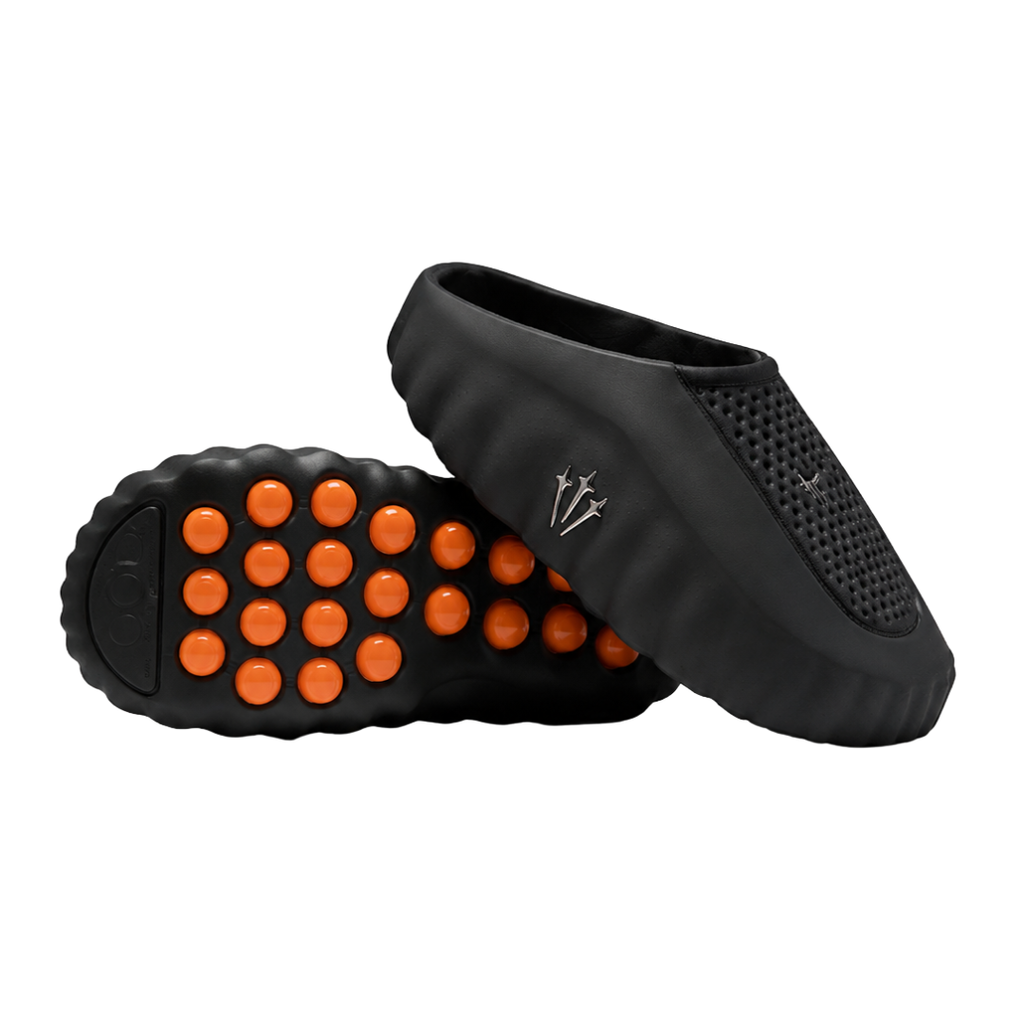 Side profile of the NOCTA x Nike Mind 001 slide, showcasing the sleek black molded upper, signature metallic star-shaped branding on the sidewall, and the vibrant orange massage-nodule outsole.
