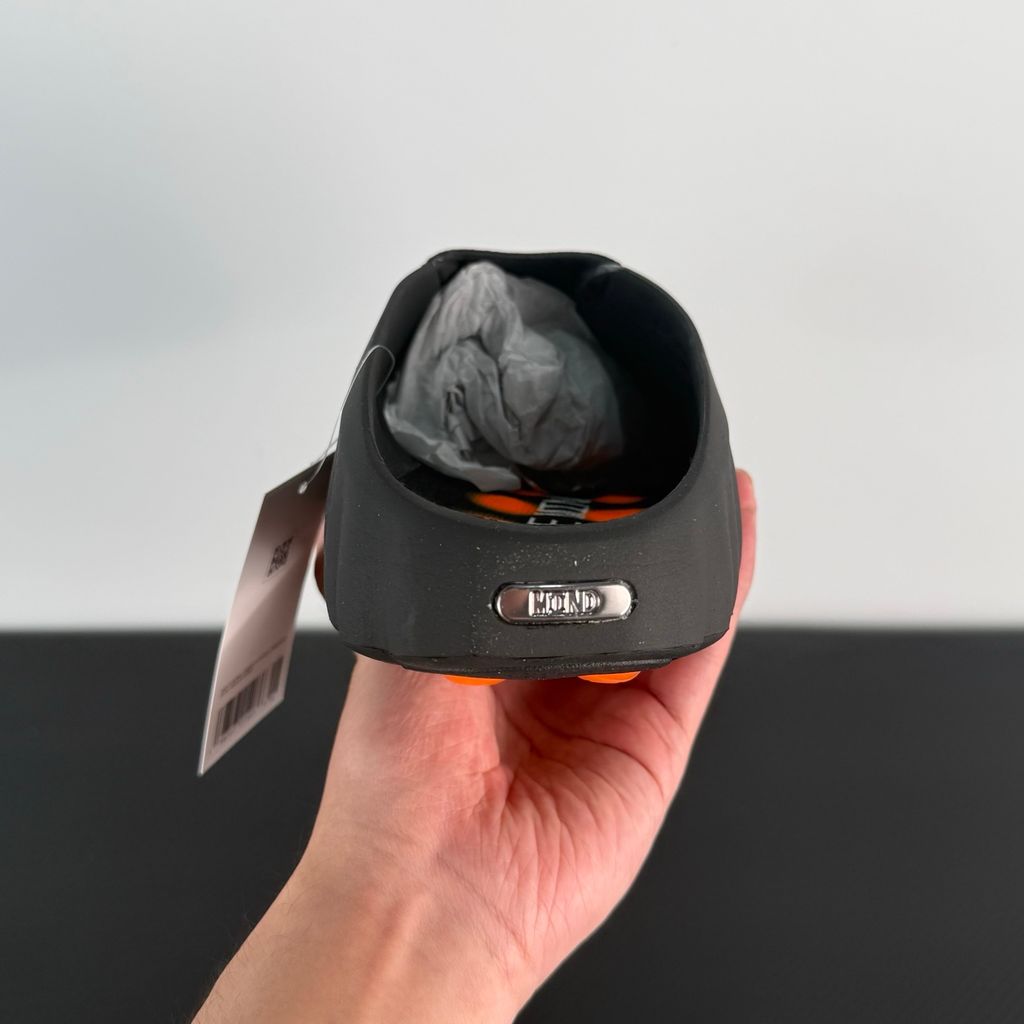 Side profile of the NOCTA x Nike Mind 001 slide, showcasing the sleek black molded upper, signature metallic star-shaped branding on the sidewall, and the vibrant orange massage-nodule outsole.