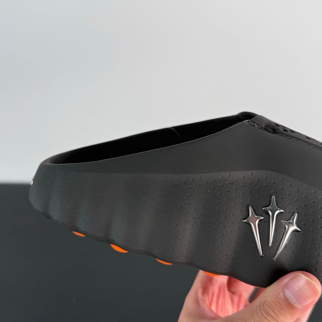 Side profile of the NOCTA x Nike Mind 001 slide, showcasing the sleek black molded upper, signature metallic star-shaped branding on the sidewall, and the vibrant orange massage-nodule outsole.
