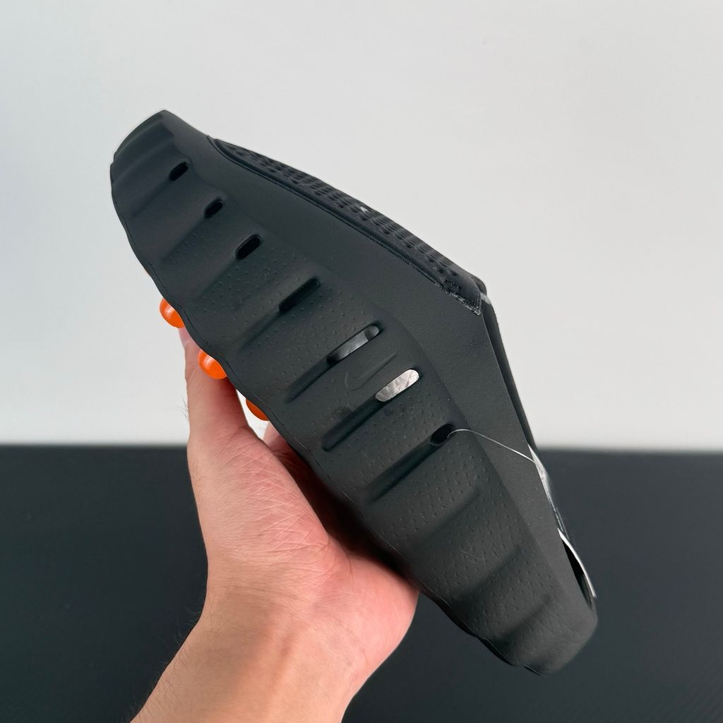 Side profile of the NOCTA x Nike Mind 001 slide, showcasing the sleek black molded upper, signature metallic star-shaped branding on the sidewall, and the vibrant orange massage-nodule outsole.