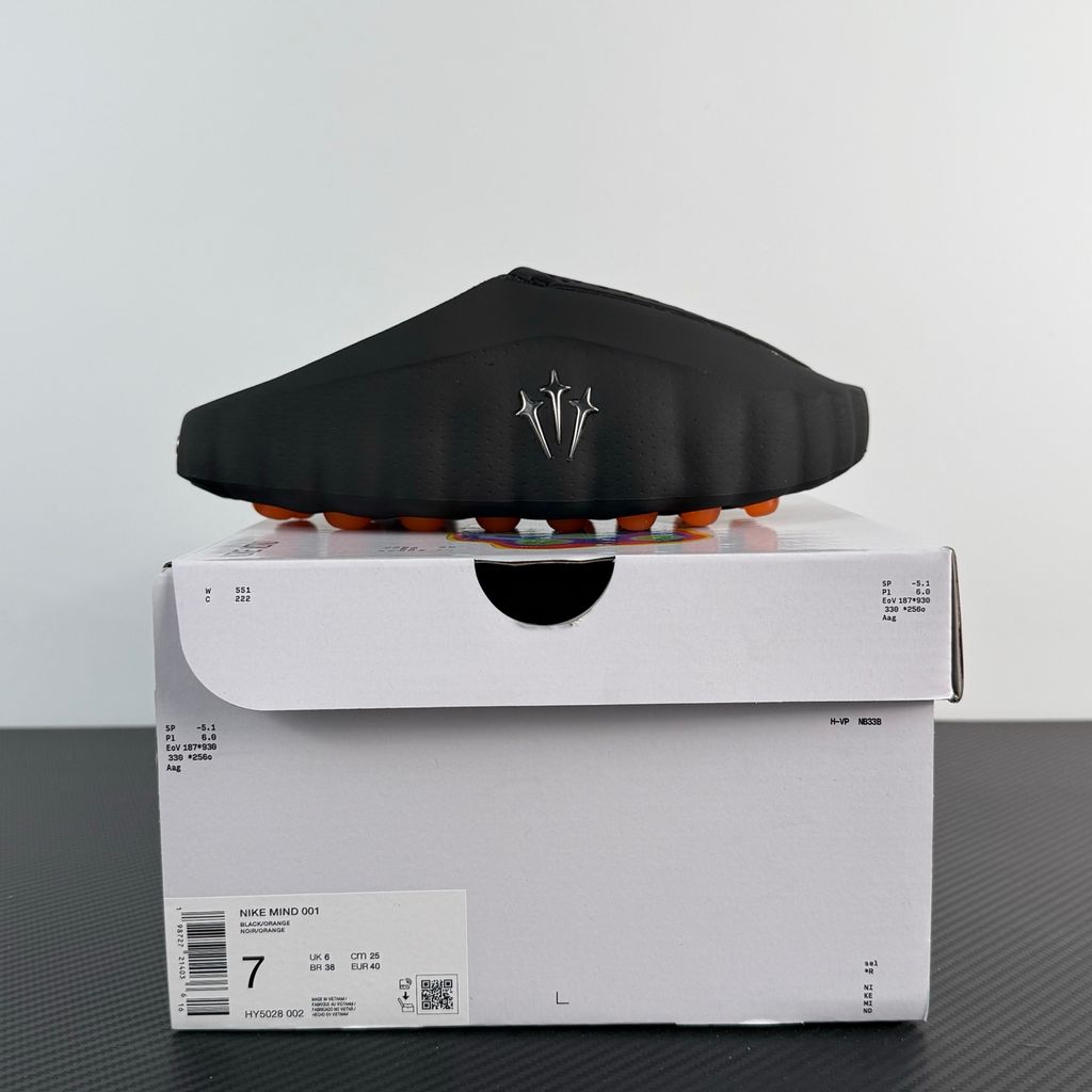 Side profile of the NOCTA x Nike Mind 001 slide, showcasing the sleek black molded upper, signature metallic star-shaped branding on the sidewall, and the vibrant orange massage-nodule outsole.