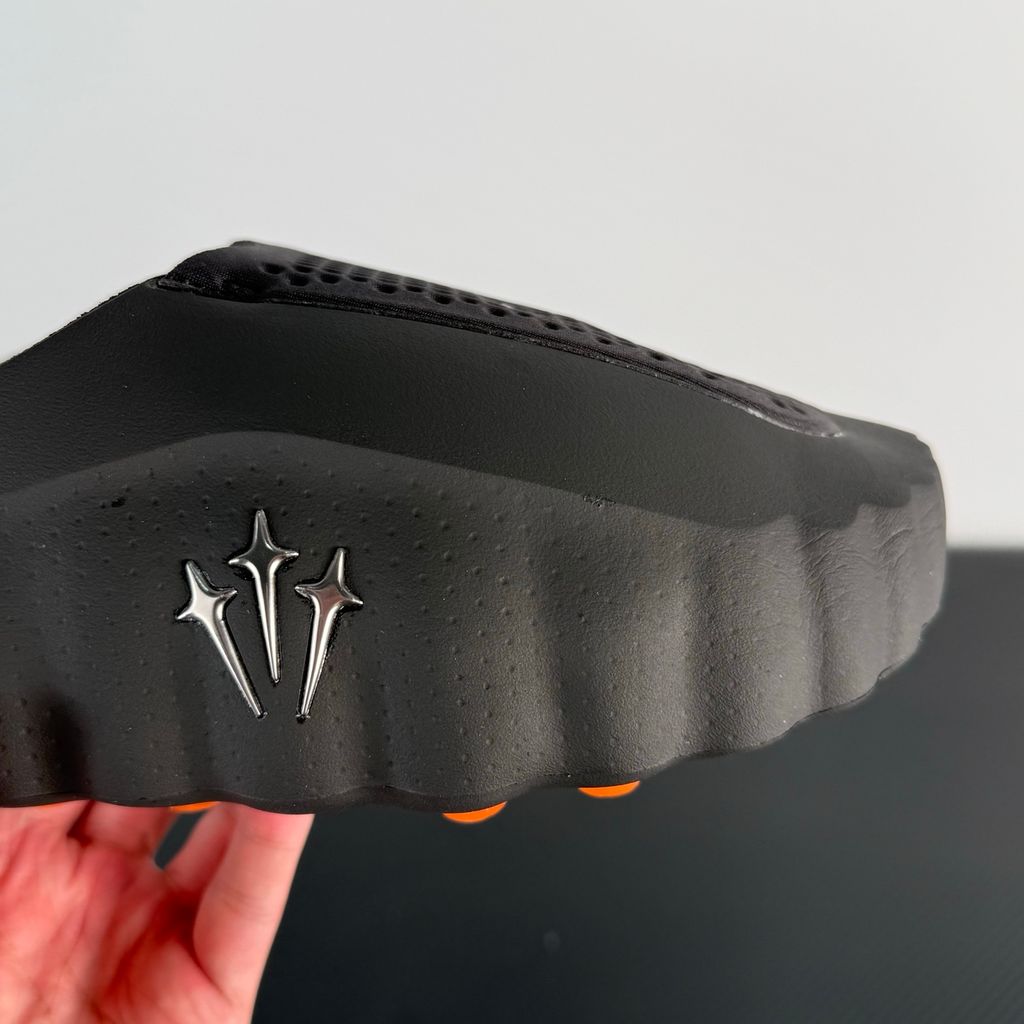 Side profile of the NOCTA x Nike Mind 001 slide, showcasing the sleek black molded upper, signature metallic star-shaped branding on the sidewall, and the vibrant orange massage-nodule outsole.