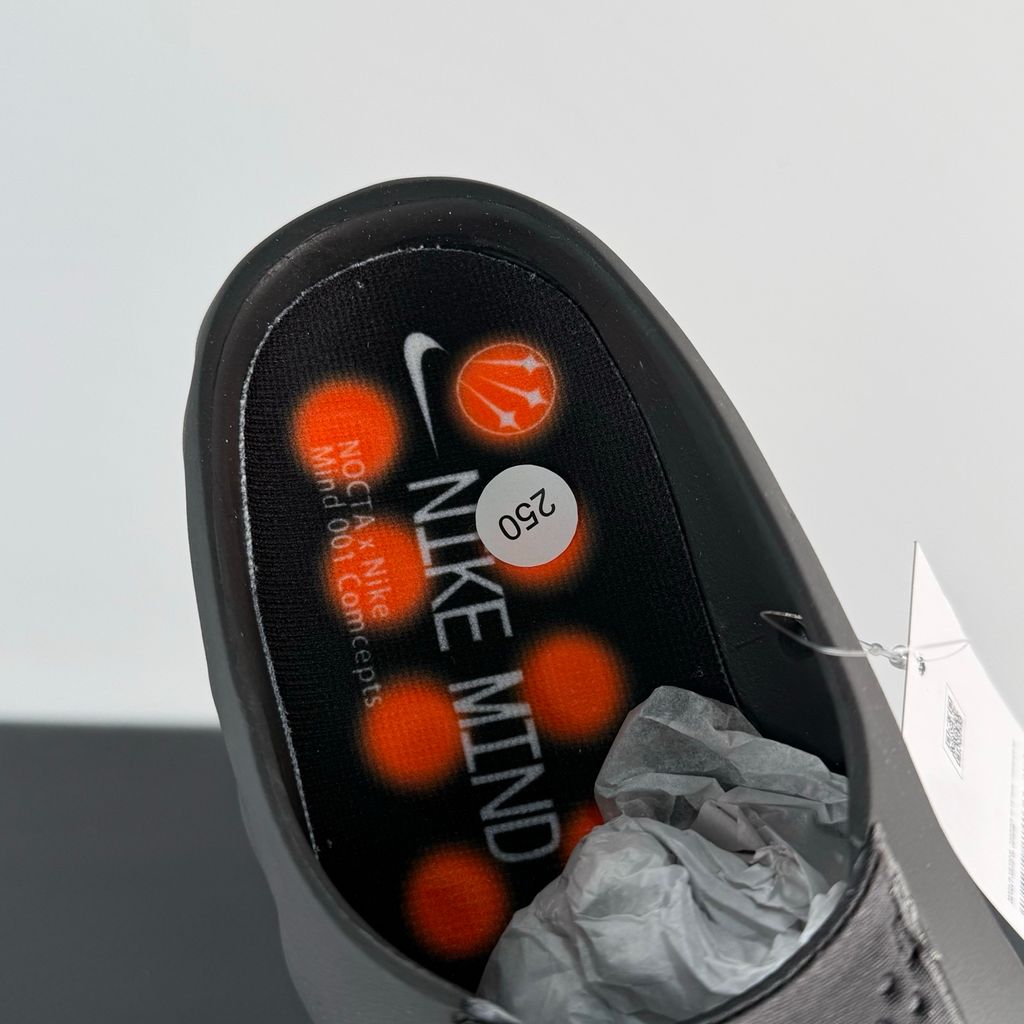 Side profile of the NOCTA x Nike Mind 001 slide, showcasing the sleek black molded upper, signature metallic star-shaped branding on the sidewall, and the vibrant orange massage-nodule outsole.