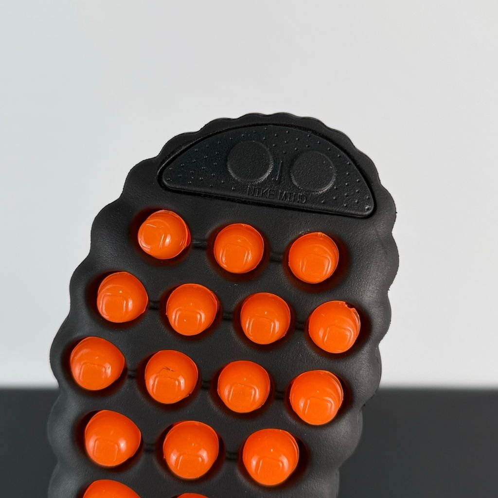Side profile of the NOCTA x Nike Mind 001 slide, showcasing the sleek black molded upper, signature metallic star-shaped branding on the sidewall, and the vibrant orange massage-nodule outsole.