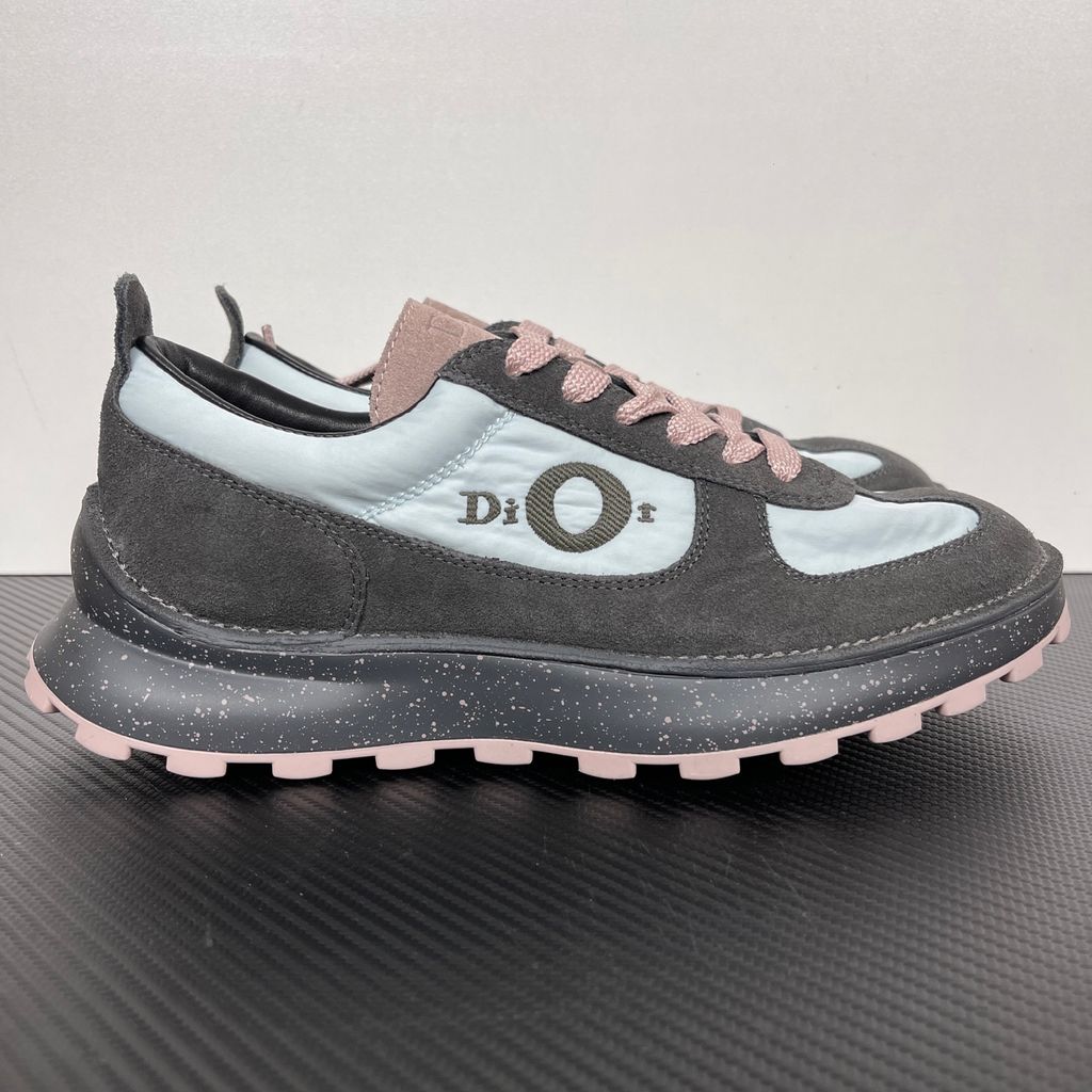 Side profile of the Dior Squash sneaker, highlighting the contrasting dark grey suede and light blue nylon upper, the unique 'DIOR' circular logo embroidery, and the speckled polyurethane midsole with a lugged outsole.