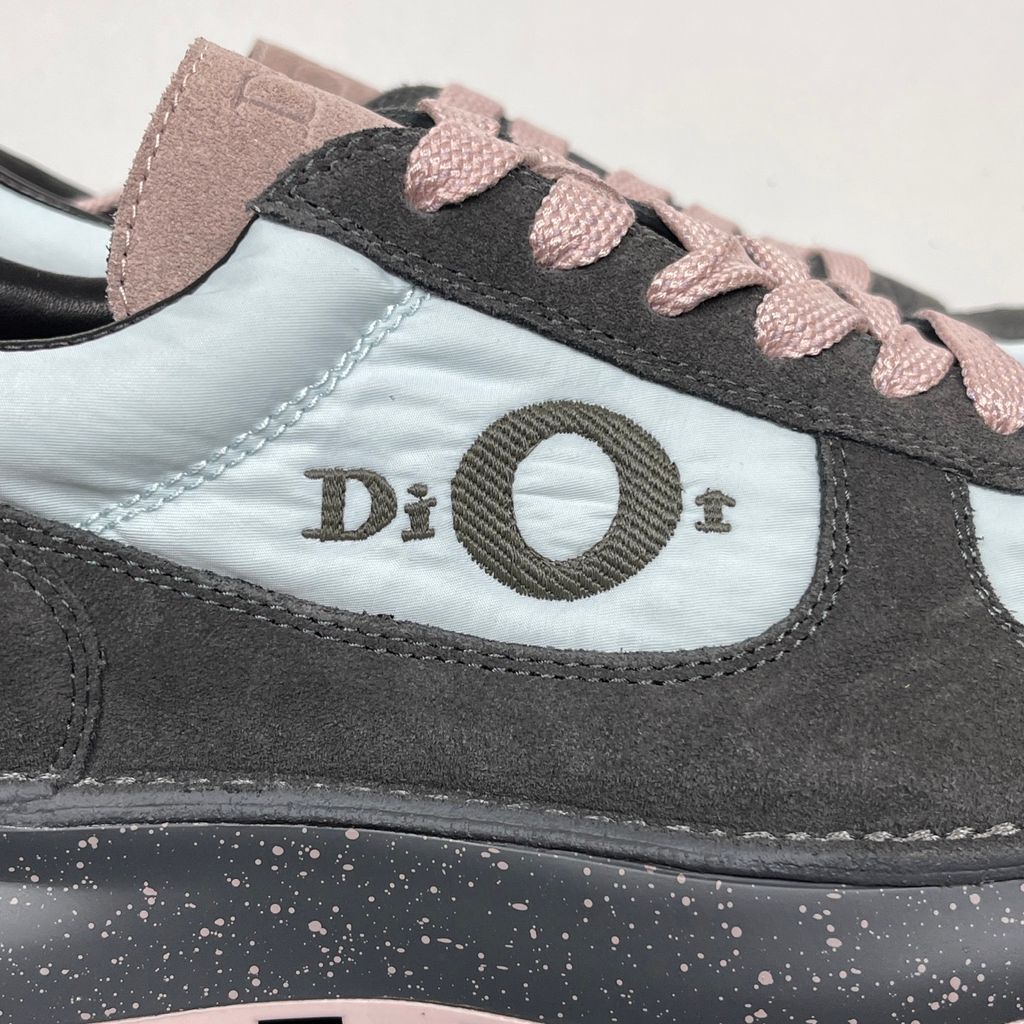 Side profile of the Dior Squash sneaker, highlighting the contrasting dark grey suede and light blue nylon upper, the unique 'DIOR' circular logo embroidery, and the speckled polyurethane midsole with a lugged outsole.