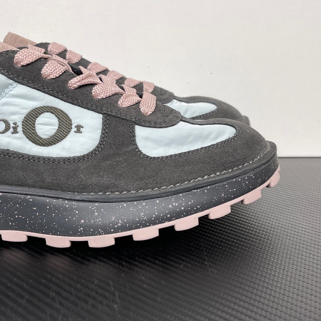Side profile of the Dior Squash sneaker, highlighting the contrasting dark grey suede and light blue nylon upper, the unique 'DIOR' circular logo embroidery, and the speckled polyurethane midsole with a lugged outsole.