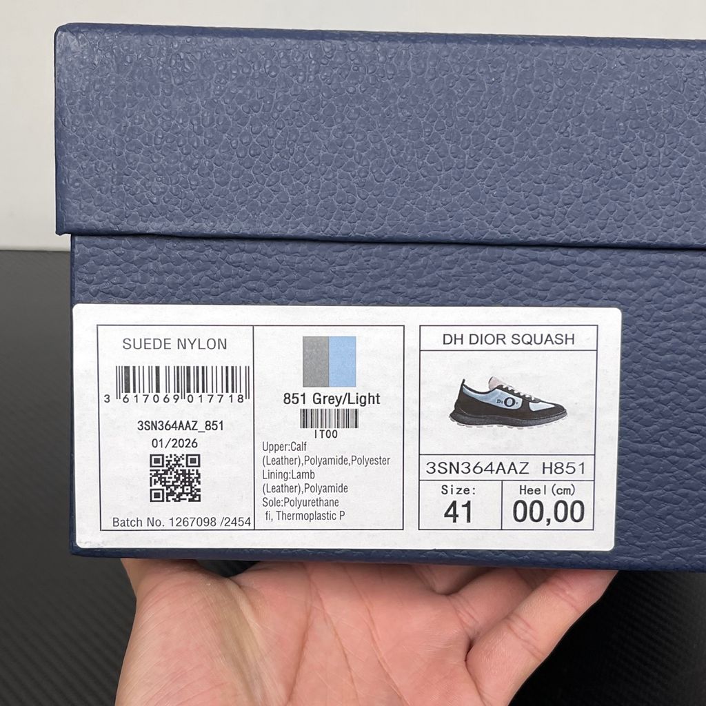 Side profile of the Dior Squash sneaker, highlighting the contrasting dark grey suede and light blue nylon upper, the unique 'DIOR' circular logo embroidery, and the speckled polyurethane midsole with a lugged outsole.