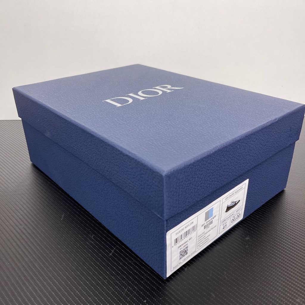 Side profile of the Dior Squash sneaker, highlighting the contrasting dark grey suede and light blue nylon upper, the unique 'DIOR' circular logo embroidery, and the speckled polyurethane midsole with a lugged outsole.