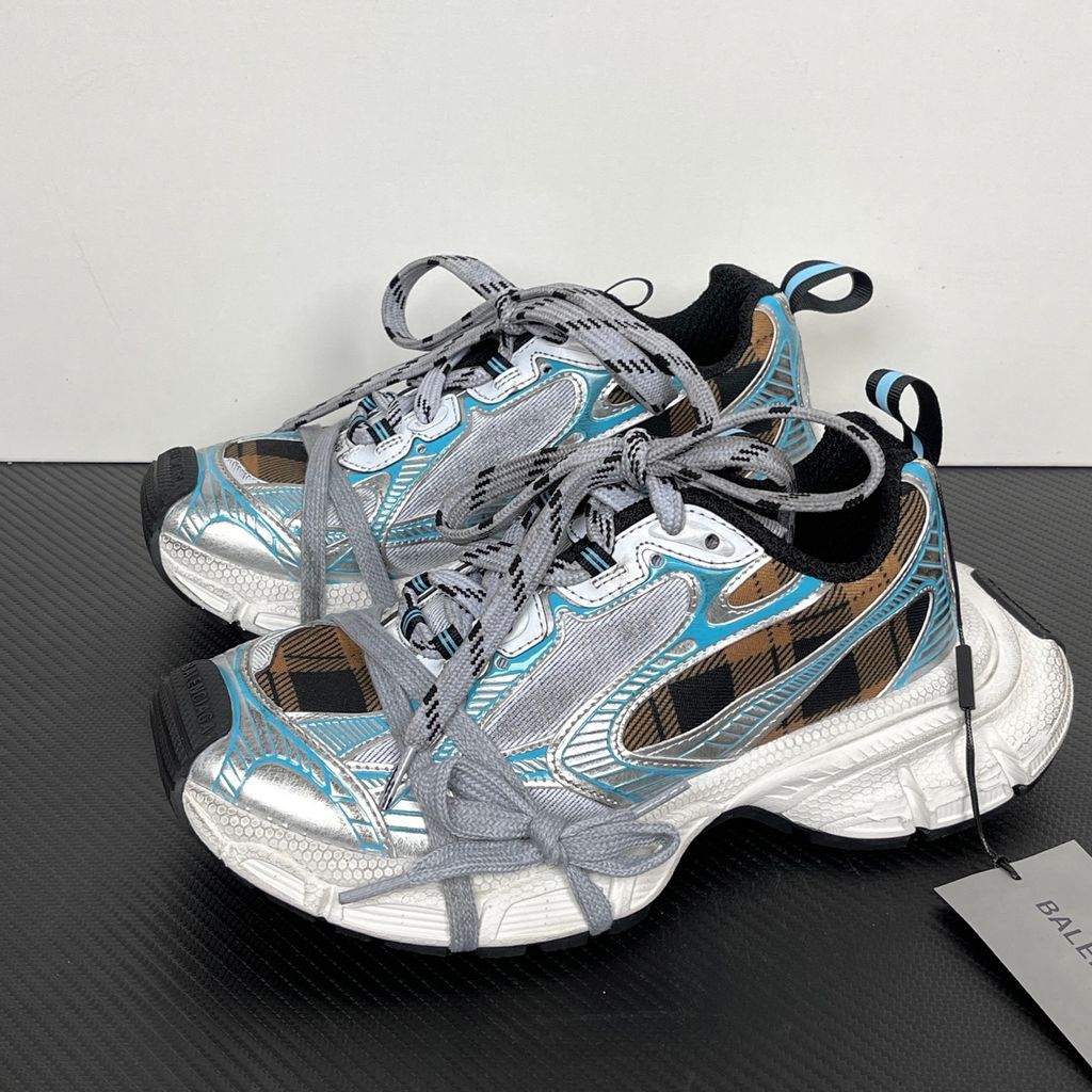 Side profile of the Balenciaga 3XL Sneaker showcasing a brown and black check pattern base, metallic silver overlays, light blue piping, a chunky distressed white sole, and the signature extra laces wrapped around the toe box.