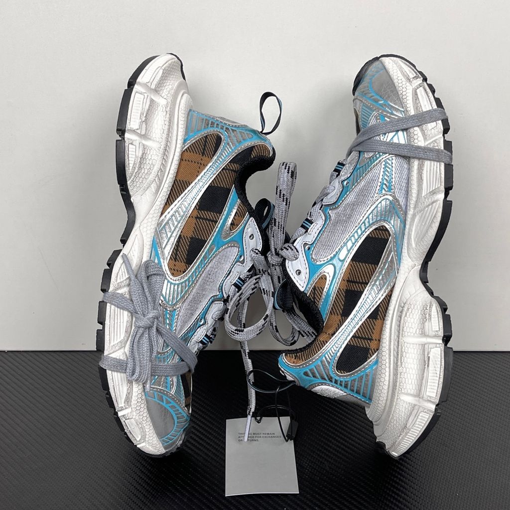 Side profile of the Balenciaga 3XL Sneaker showcasing a brown and black check pattern base, metallic silver overlays, light blue piping, a chunky distressed white sole, and the signature extra laces wrapped around the toe box.