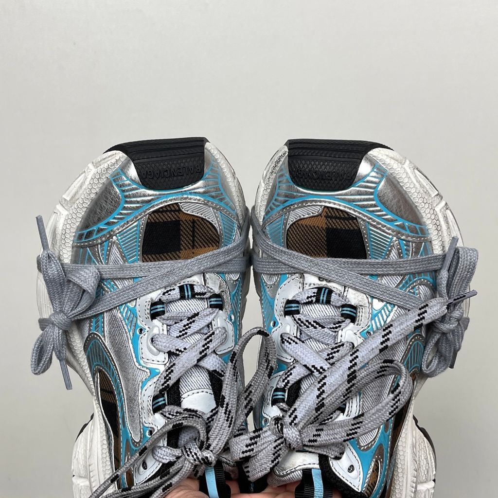 Side profile of the Balenciaga 3XL Sneaker showcasing a brown and black check pattern base, metallic silver overlays, light blue piping, a chunky distressed white sole, and the signature extra laces wrapped around the toe box.