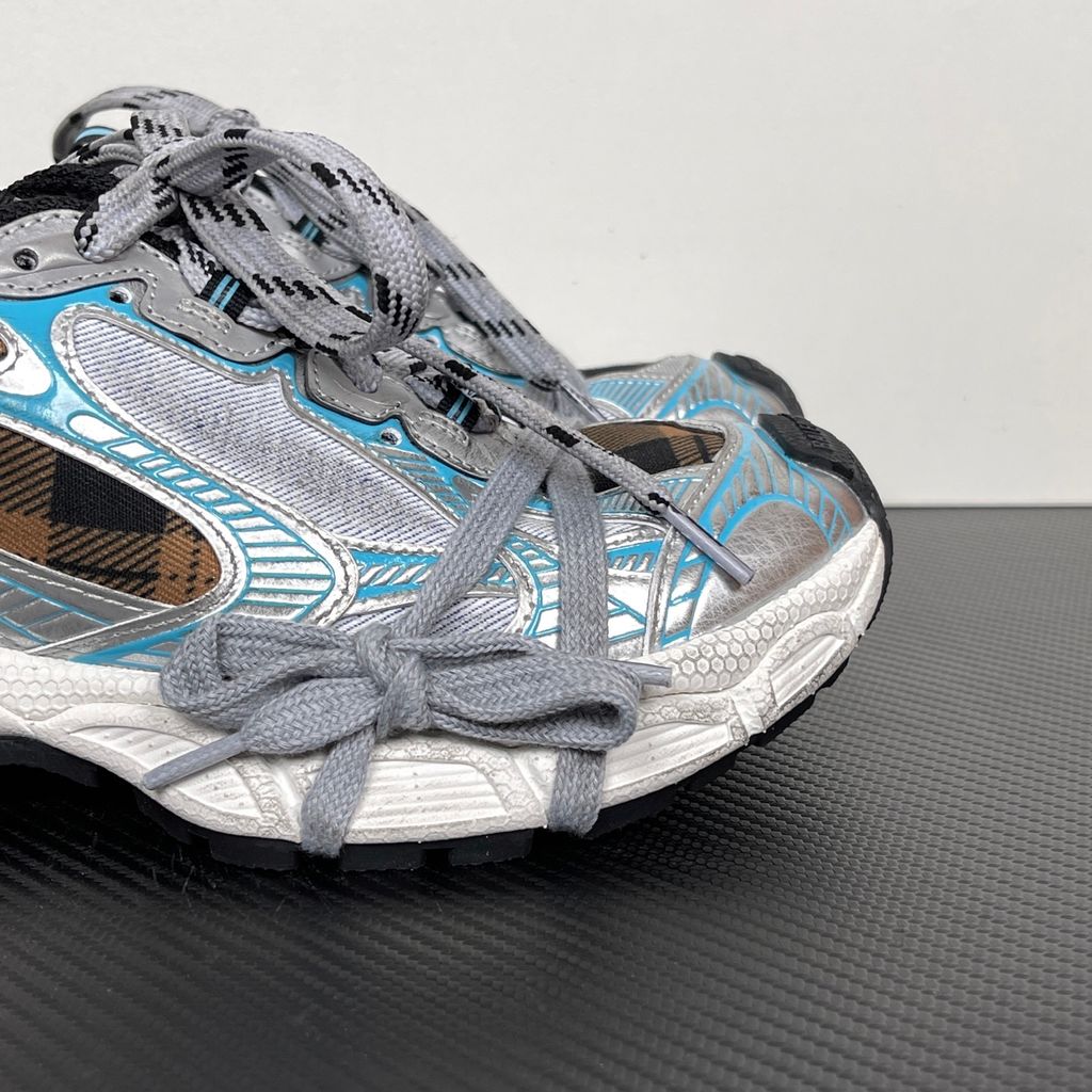 Side profile of the Balenciaga 3XL Sneaker showcasing a brown and black check pattern base, metallic silver overlays, light blue piping, a chunky distressed white sole, and the signature extra laces wrapped around the toe box.