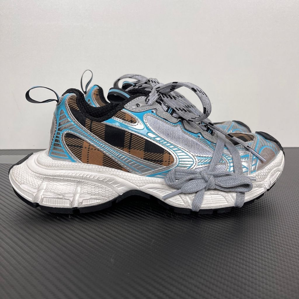 Side profile of the Balenciaga 3XL Sneaker showcasing a brown and black check pattern base, metallic silver overlays, light blue piping, a chunky distressed white sole, and the signature extra laces wrapped around the toe box.