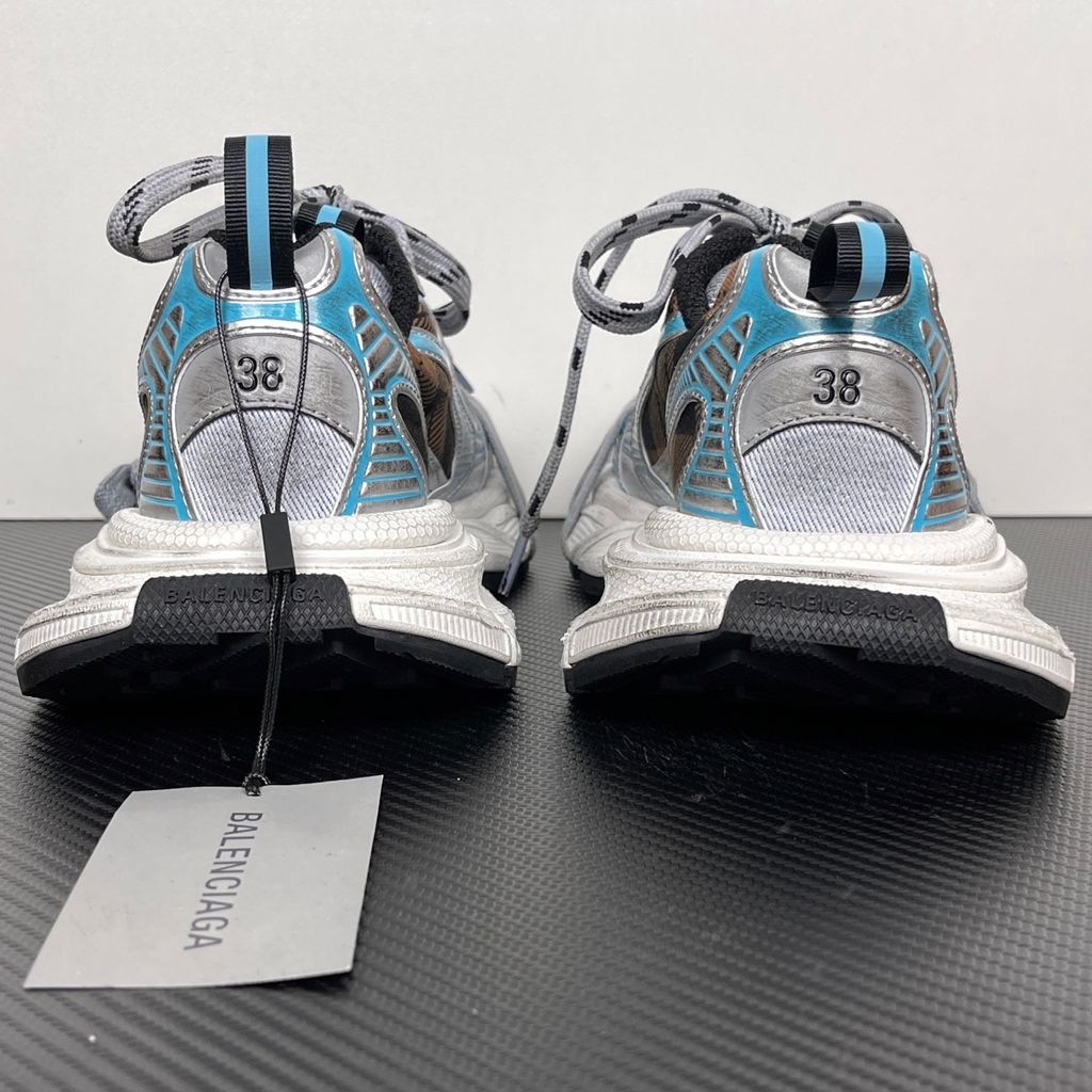 Side profile of the Balenciaga 3XL Sneaker showcasing a brown and black check pattern base, metallic silver overlays, light blue piping, a chunky distressed white sole, and the signature extra laces wrapped around the toe box.