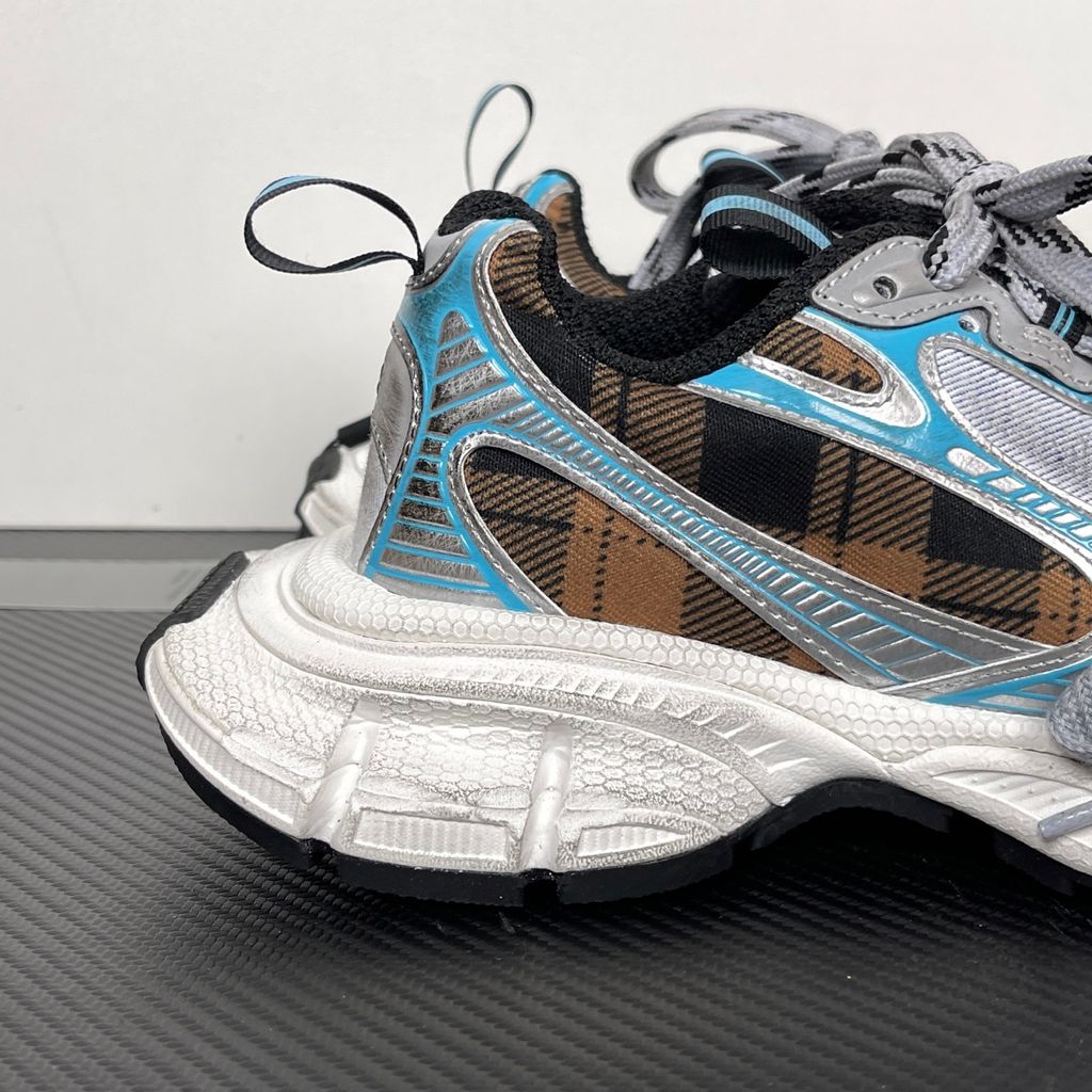 Side profile of the Balenciaga 3XL Sneaker showcasing a brown and black check pattern base, metallic silver overlays, light blue piping, a chunky distressed white sole, and the signature extra laces wrapped around the toe box.