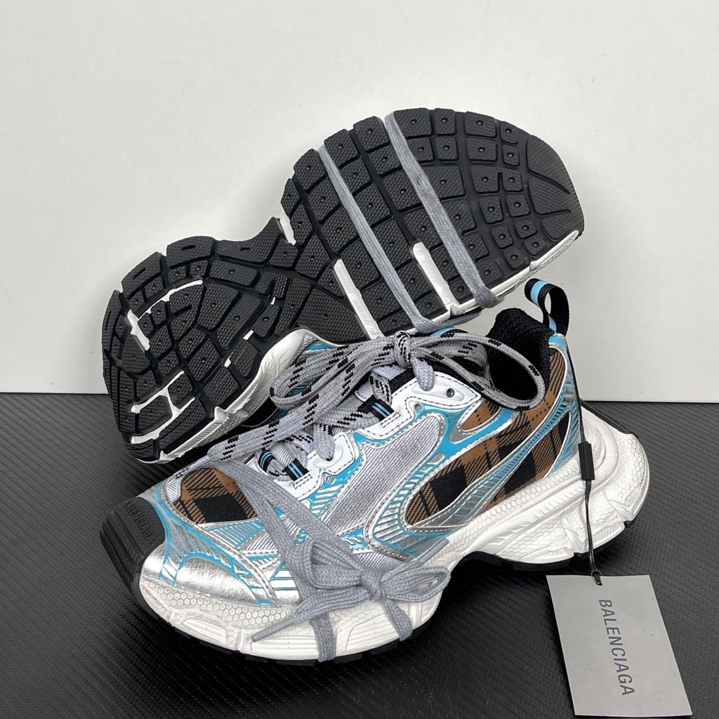 Side profile of the Balenciaga 3XL Sneaker showcasing a brown and black check pattern base, metallic silver overlays, light blue piping, a chunky distressed white sole, and the signature extra laces wrapped around the toe box.