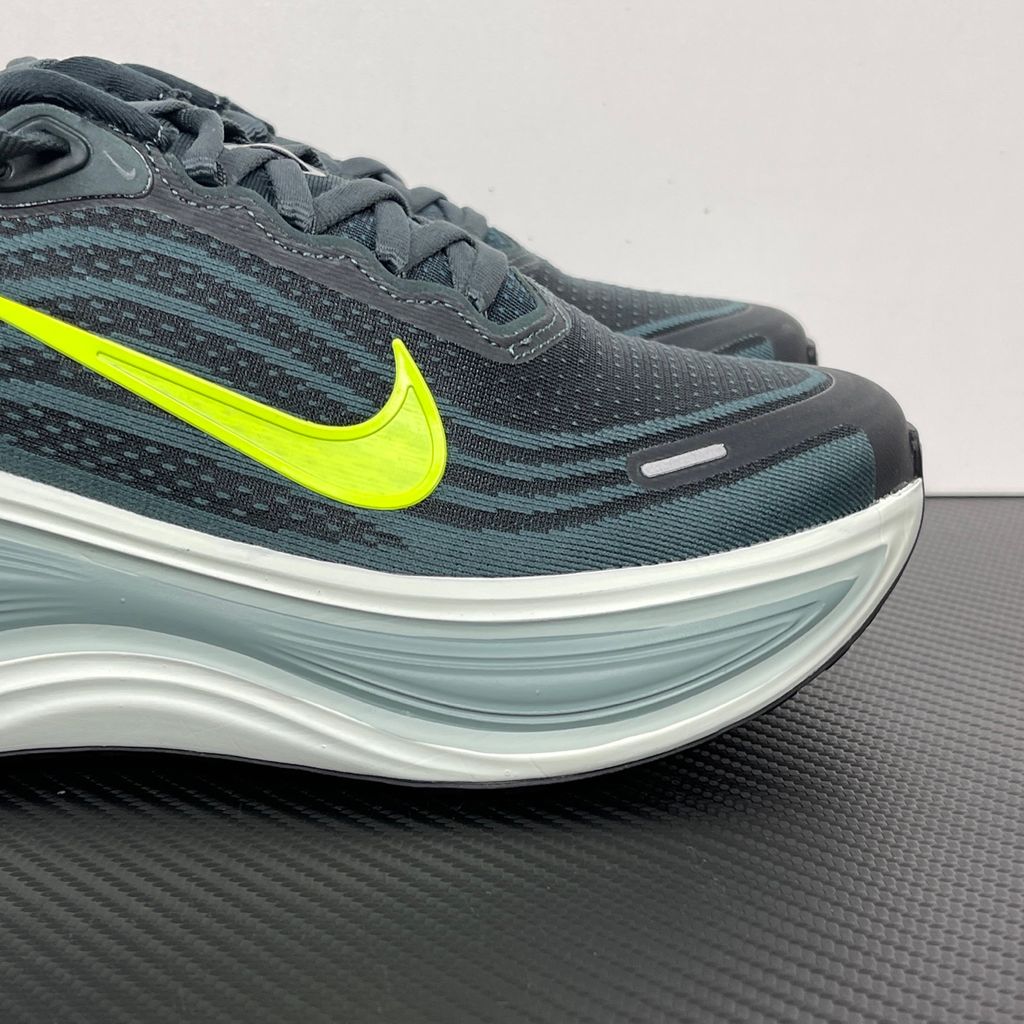 Side profile of the Nike Vomero Plus running shoe, featuring an earthy green engineered mesh upper with Seaweed and Cannon overlays, striking neon Volt accents, and a heavily cushioned, sculpted ZoomX foam midsole.