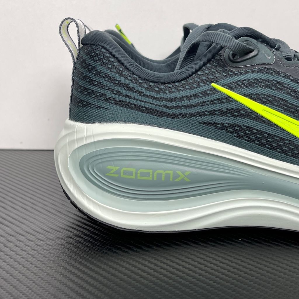 Side profile of the Nike Vomero Plus running shoe, featuring an earthy green engineered mesh upper with Seaweed and Cannon overlays, striking neon Volt accents, and a heavily cushioned, sculpted ZoomX foam midsole.