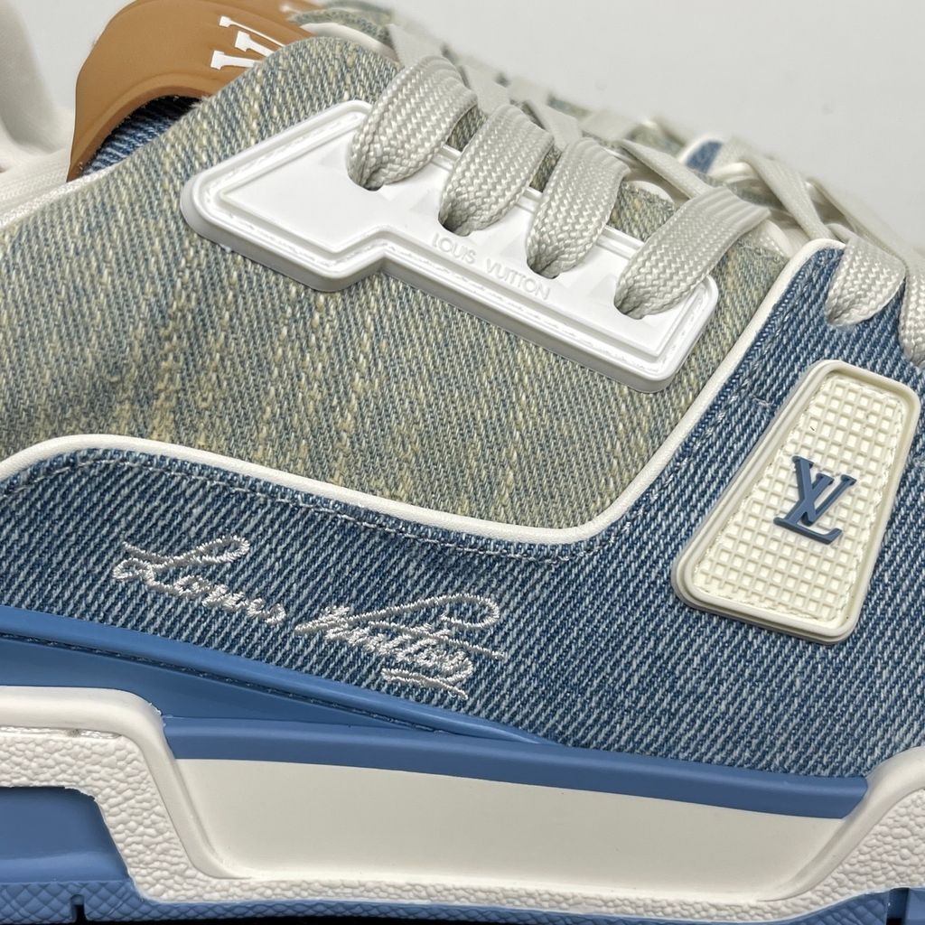 Side profile of the Tyler, the Creator x Louis Vuitton LV Trainer sneaker, showcasing a mix of light grey and blue denim paneling, a contrasting brown leather heel tab, the LV Initials logo, and custom cursive Louis Vuitton embroidery on the lateral side.