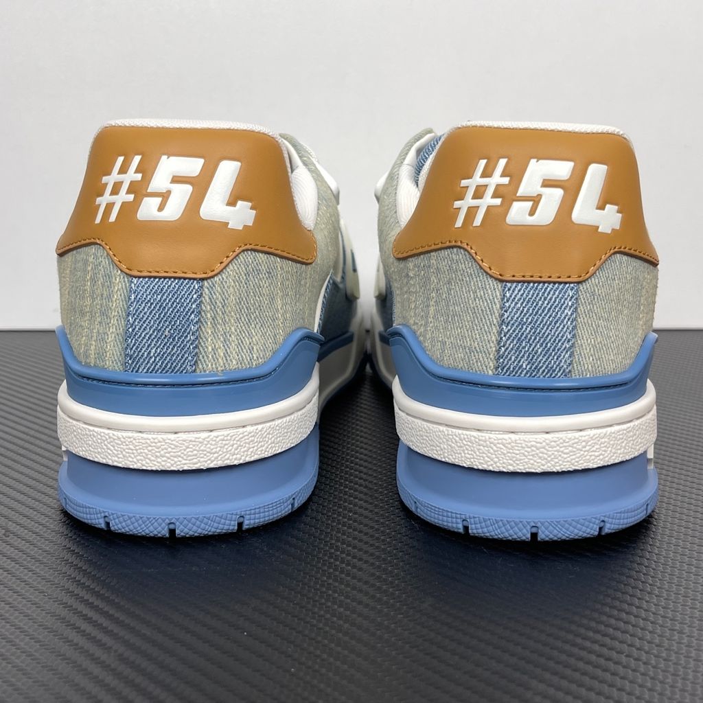 Side profile of the Tyler, the Creator x Louis Vuitton LV Trainer sneaker, showcasing a mix of light grey and blue denim paneling, a contrasting brown leather heel tab, the LV Initials logo, and custom cursive Louis Vuitton embroidery on the lateral side.