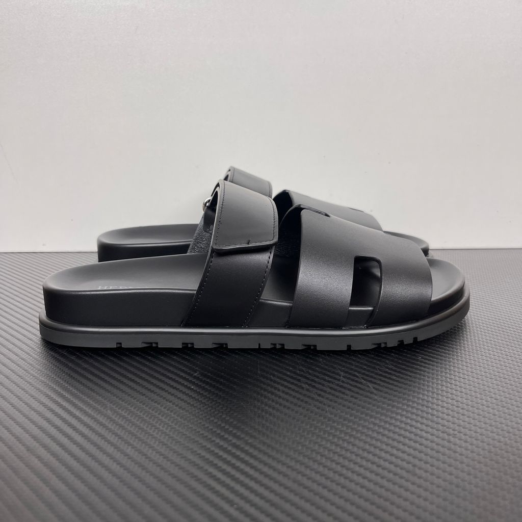Side profile of the Hermès Chypre sandal crafted in smooth black calfskin leather, featuring the signature 'H' cut-out design across the toe, an adjustable velcro strap over the instep, and a contoured anatomical rubber sole.