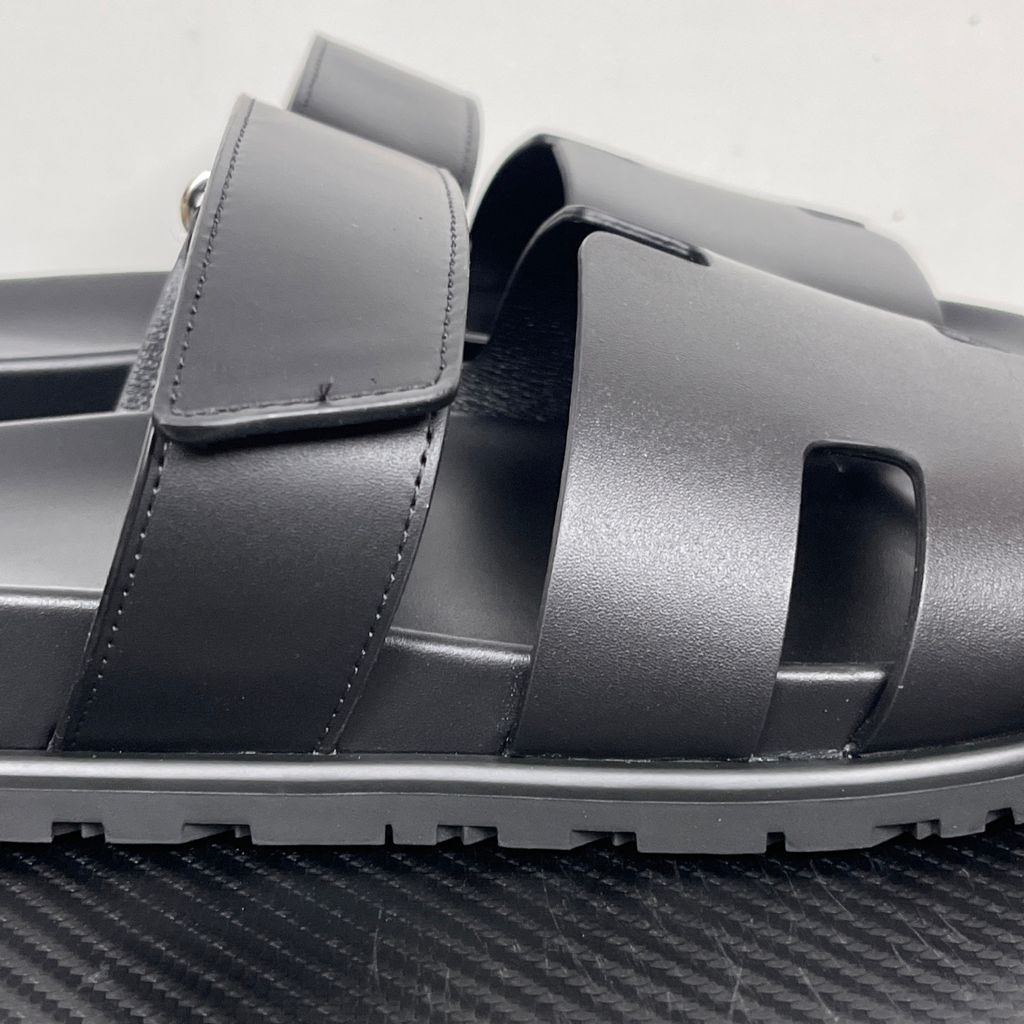Side profile of the Hermès Chypre sandal crafted in smooth black calfskin leather, featuring the signature 'H' cut-out design across the toe, an adjustable velcro strap over the instep, and a contoured anatomical rubber sole.