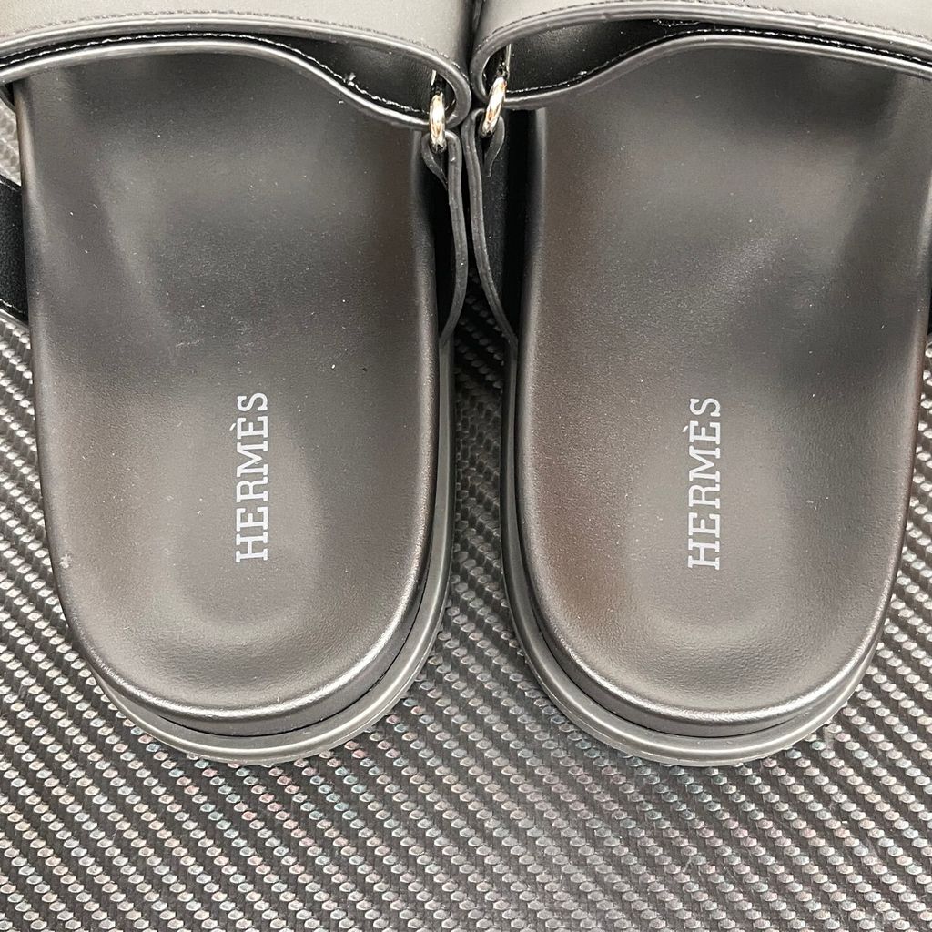 Side profile of the Hermès Chypre sandal crafted in smooth black calfskin leather, featuring the signature 'H' cut-out design across the toe, an adjustable velcro strap over the instep, and a contoured anatomical rubber sole.