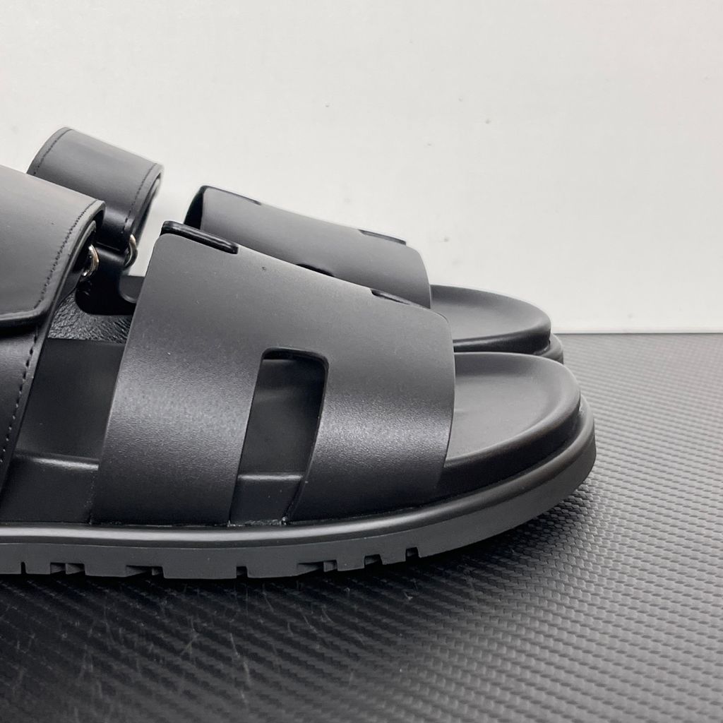 Side profile of the Hermès Chypre sandal crafted in smooth black calfskin leather, featuring the signature 'H' cut-out design across the toe, an adjustable velcro strap over the instep, and a contoured anatomical rubber sole.