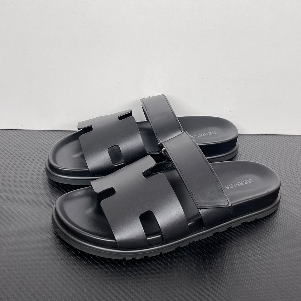 Side profile of the Hermès Chypre sandal crafted in smooth black calfskin leather, featuring the signature 'H' cut-out design across the toe, an adjustable velcro strap over the instep, and a contoured anatomical rubber sole.
