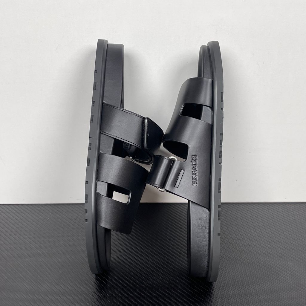 Side profile of the Hermès Chypre sandal crafted in smooth black calfskin leather, featuring the signature 'H' cut-out design across the toe, an adjustable velcro strap over the instep, and a contoured anatomical rubber sole.