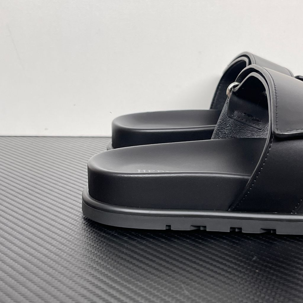 Side profile of the Hermès Chypre sandal crafted in smooth black calfskin leather, featuring the signature 'H' cut-out design across the toe, an adjustable velcro strap over the instep, and a contoured anatomical rubber sole.
