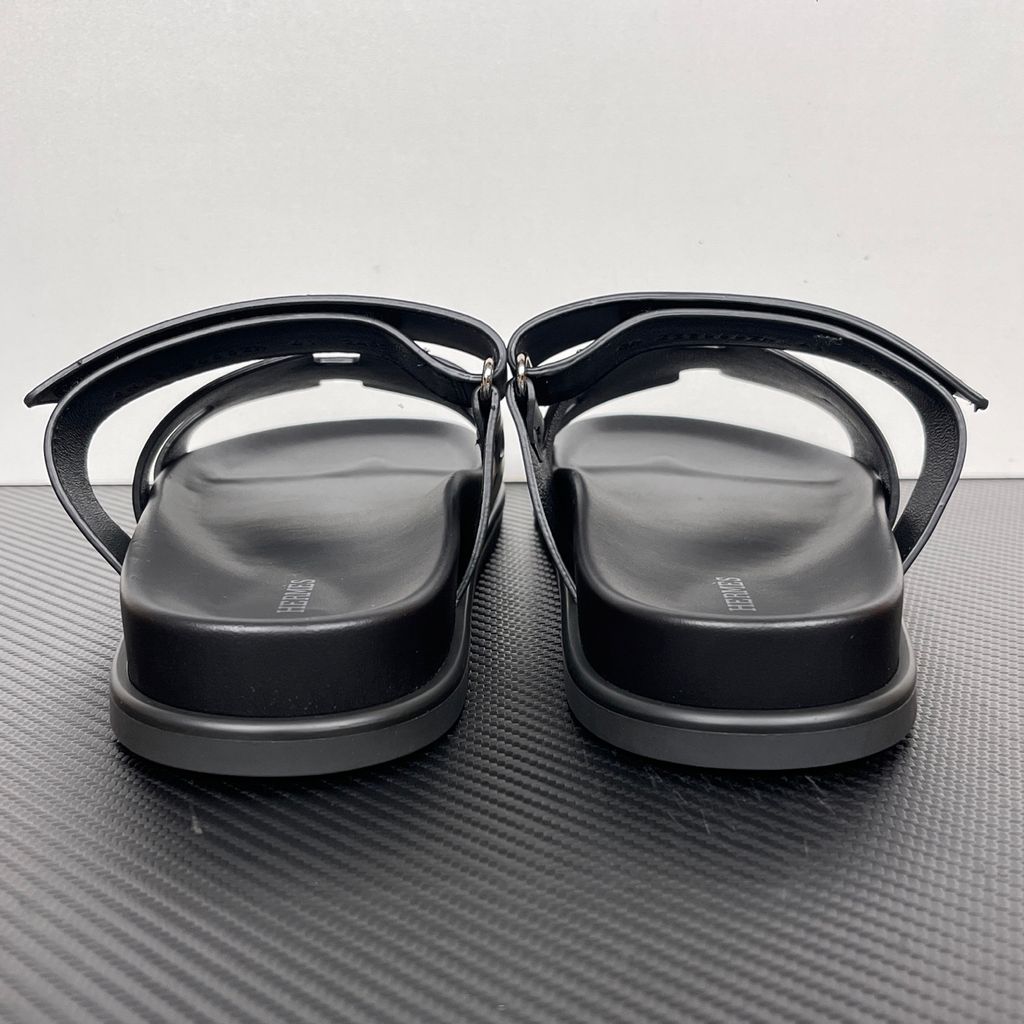 Side profile of the Hermès Chypre sandal crafted in smooth black calfskin leather, featuring the signature 'H' cut-out design across the toe, an adjustable velcro strap over the instep, and a contoured anatomical rubber sole.